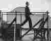 black and white image of a man in early 1900s formal clothes is walking on a huge agricultural-looking treadmill