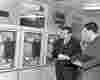 two men in suits inspect a ticker-tape read out from a 60s reel-to-reel computer