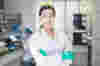 A smiling, confident female scientist wearing a lab coat stands in a lab facing the camera